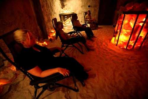 Four people relax in reclining chairs inside a dimly lit salt therapy room with glowing salt rocks.