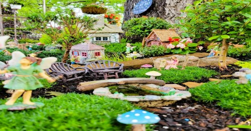 A whimsical miniature garden with fairy figurines, tiny houses, benches, and colorful plants among lush greenery.