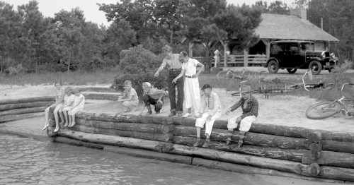 A group of children and adults sit on a log structure by a lake, with a cabin and vintage car in the background.