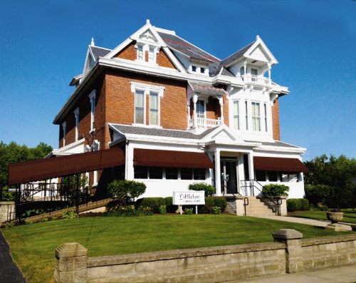 A large, elegant brick house with a white trim, featuring a porch and awnings, set in a well-maintained landscape.