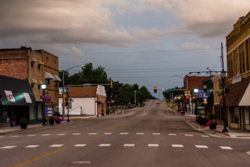 A quiet small-town street with shops, traffic lights, and cloudy skies, creating a calm, empty atmosphere.