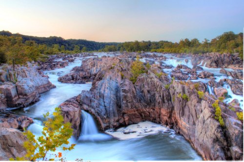 A serene river flows over rocky terrain, surrounded by lush greenery and a soft sunset glow in the background.