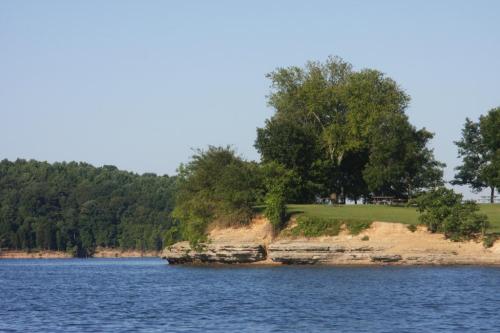 A serene lakeside view featuring a grassy shore with trees and calm blue water under a clear sky.