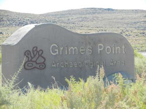 Sign for Grimes Point Archaeological Area, featuring a stylized design and surrounded by desert vegetation.