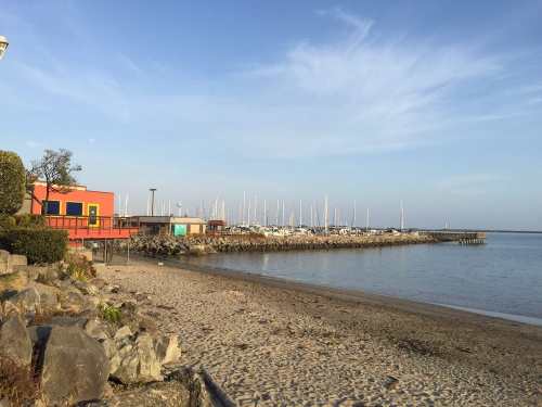 A sandy beach with a calm water view, boats docked in the distance, and colorful buildings along the shore.