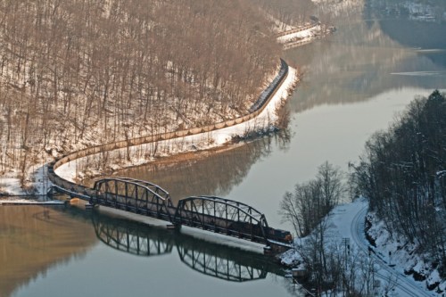 A train bridge spans a river, surrounded by snow-covered trees and a winding path along the water's edge.