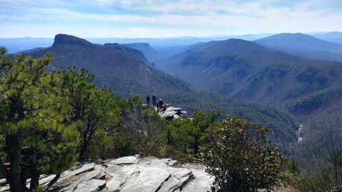 A scenic mountain view with hikers on a rocky ledge, surrounded by lush greenery and distant blue peaks under a clear sky.