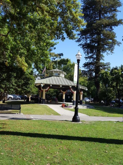 A park gazebo surrounded by trees, benches, and a lamp post on a sunny day.