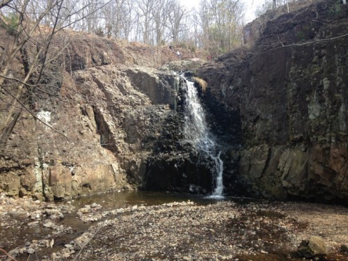 A waterfall cascading down rocky cliffs into a shallow stream, surrounded by bare trees and rocky terrain.