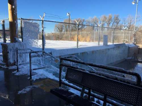 A frozen outdoor area with a bench, icy fence, and snow-covered ground under a clear blue sky.