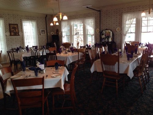 A cozy dining room with tables set for a meal, featuring purple napkins and wooden chairs in a charming, vintage style.