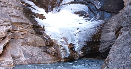 A frozen waterfall cascading down rocky cliffs into a small pool of water below. Ice and snow cover the rocks.