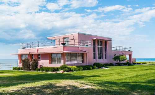 A pink modern house with large windows, surrounded by green grass and overlooking a body of water under a blue sky.