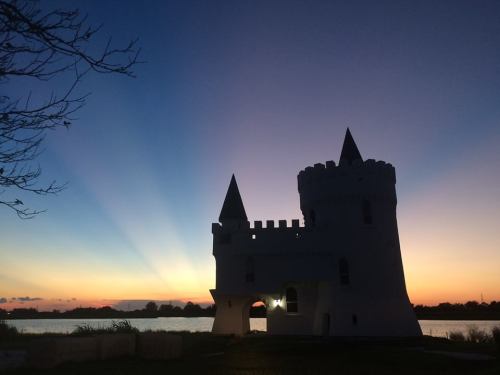 Silhouette of a castle against a colorful sunset, with rays of light illuminating the sky over a calm water body.