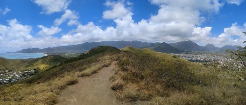 A panoramic view of a grassy hillside with mountains and a coastal town under a partly cloudy sky.