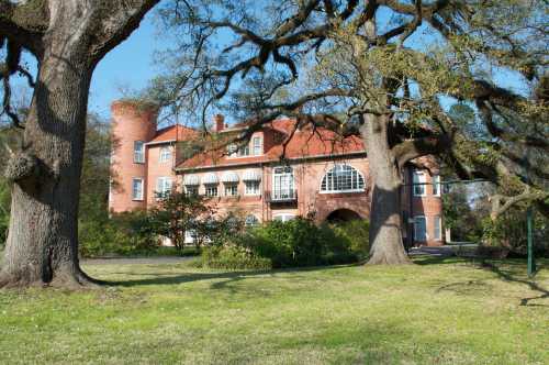 A large brick house with a red roof, surrounded by trees and greenery on a sunny day.