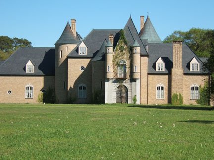 A large, castle-like brick mansion with turrets, set in a green field under a clear blue sky.