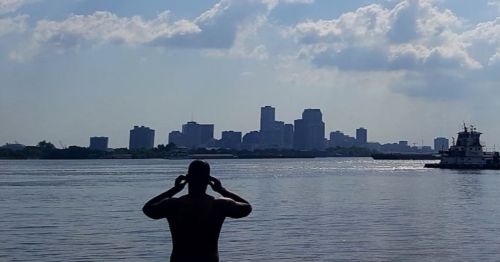 A person stands by the water, looking at a city skyline under a cloudy sky. A boat is visible nearby.