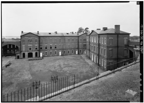 Historic brick buildings surrounding a grassy courtyard, with a wrought iron fence in the foreground. Overcast sky.