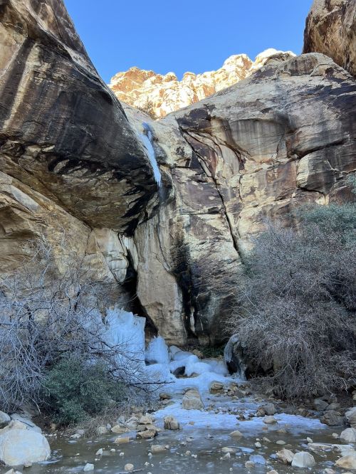 A frozen waterfall cascades down rocky cliffs, surrounded by sparse vegetation and a clear blue sky.