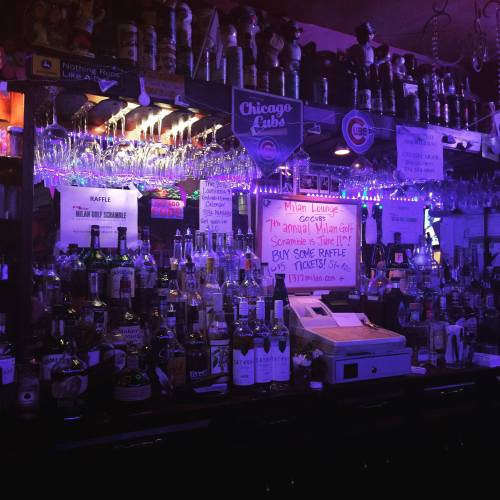 A dimly lit bar with colorful lights, shelves filled with liquor bottles, and a sign for a raffle event.