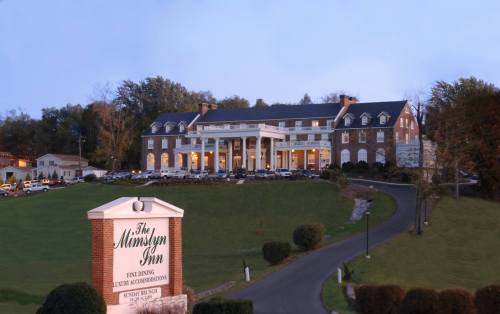 A large, elegant hotel with a white-columned entrance, set on a green hillside at dusk. The sign reads "The Mindylyn Inn."