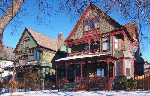 Two Victorian-style houses with intricate woodwork, one green and one red, set against a clear blue sky. Snow covers the ground.
