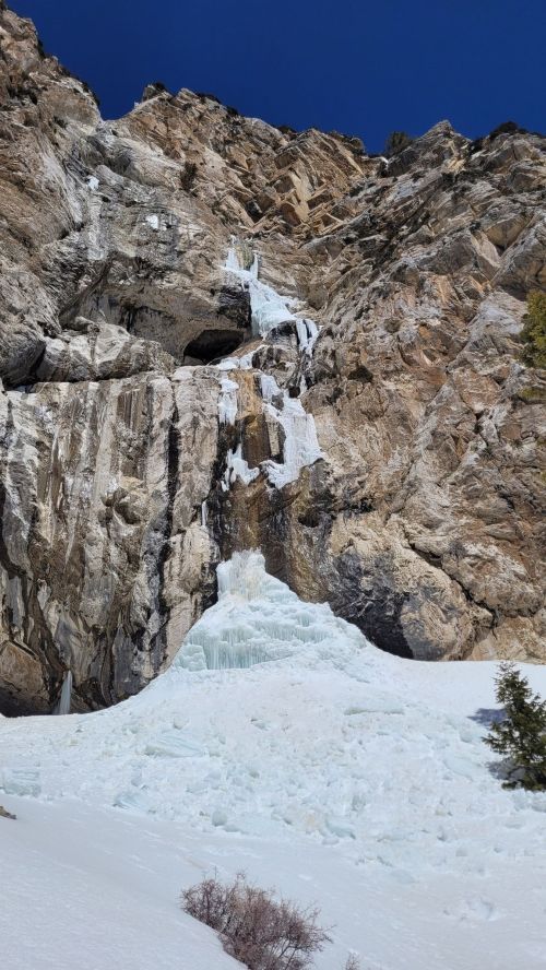 A frozen waterfall cascading down a rocky cliff, surrounded by snow and a clear blue sky.