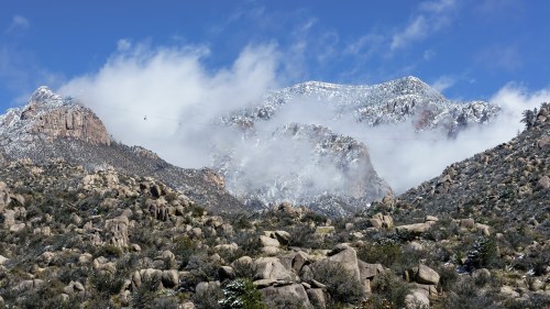 Snow-capped mountains rise above rocky terrain, partially shrouded in clouds under a bright blue sky.