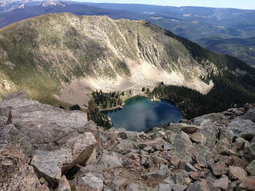 A scenic view from a rocky mountain peak overlooking a tranquil lake surrounded by forested hills.