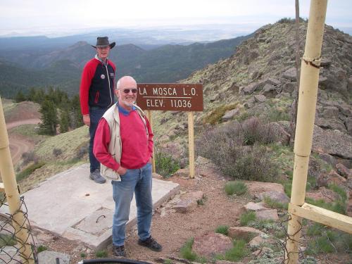Two men stand at a mountain lookout sign reading "La Mosca L.O. Elev. 11036," with a scenic view in the background.