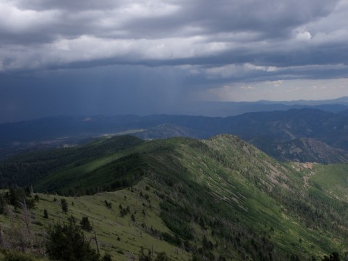 A panoramic view of green mountains under a cloudy sky, with dark rain clouds in the distance.