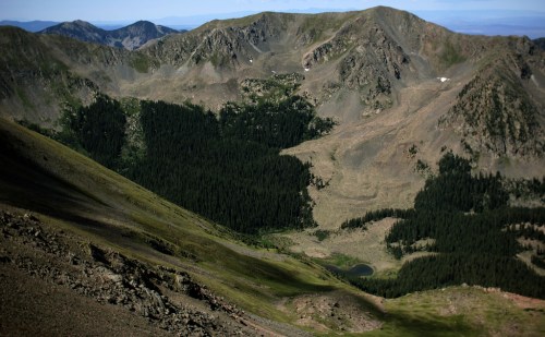 A panoramic view of rugged mountains with a forested valley and a small pond nestled among the peaks.