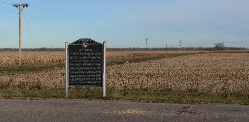 A historical marker stands beside a barren field, with power lines stretching into the distance under a clear sky.