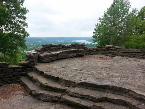 A stone observation platform surrounded by trees, overlooking a valley and river under a cloudy sky.