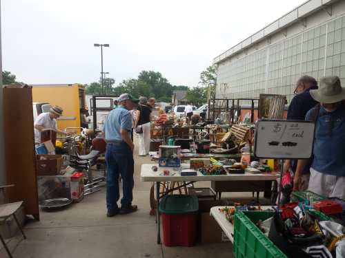 A bustling outdoor flea market with various tables of items for sale and shoppers browsing among the goods.