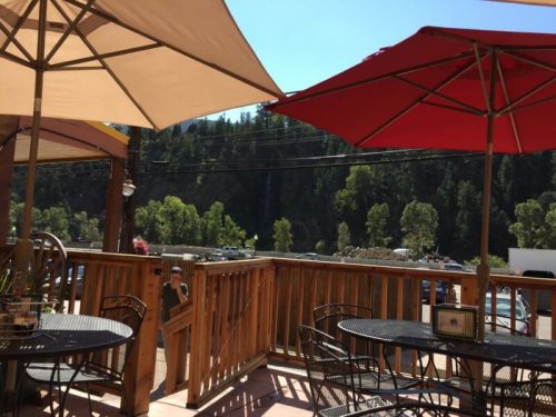 Outdoor dining area with black tables and umbrellas, overlooking a scenic view of trees and a parking area.