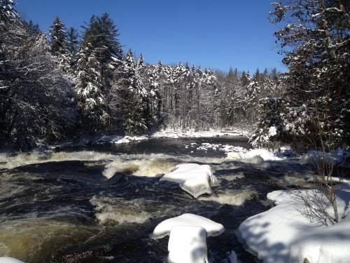 A snowy river scene with frosty trees and flowing water under a clear blue sky.