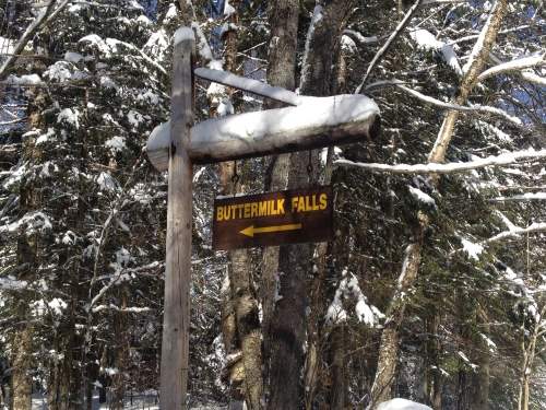 Wooden sign pointing left to Buttermilk Falls, surrounded by snow-covered trees.