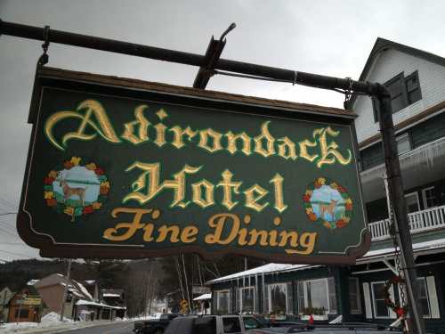 Sign for Adirondack Hotel featuring decorative text and floral accents, with a snowy building in the background.