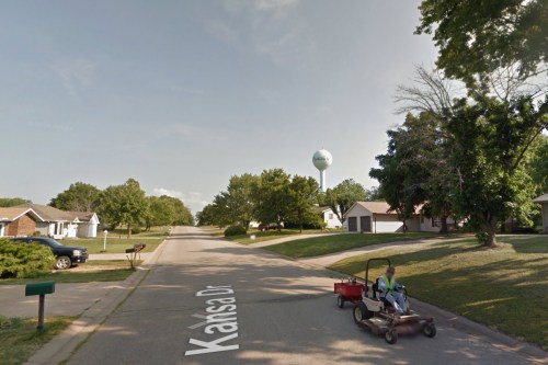 A person on a riding lawn mower drives down Kansas Dr, with a water tower and houses in the background.