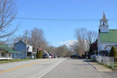 A quiet rural street lined with houses and a church, under a clear blue sky with a few trees and American flags.