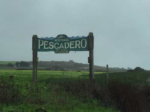 Sign for "Historic Pescadero" with a green and blue design, set against a foggy landscape and grassy fields.