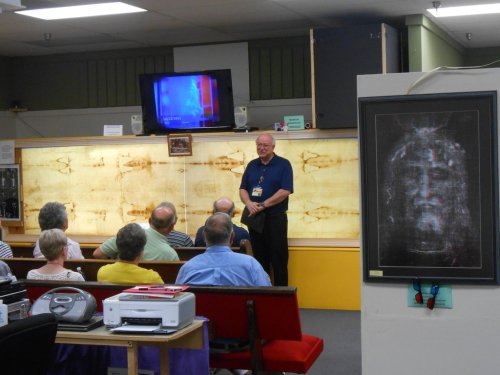 A speaker presents to an audience in a room featuring a large display of the Shroud of Turin.