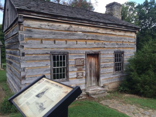 A rustic log cabin with a stone chimney, surrounded by greenery and a historical information sign in the foreground.