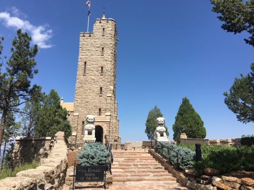 A stone tower with a flag, flanked by lion statues, surrounded by trees and blue sky. Sign reads "Will Rogers Shrine of the Sun."