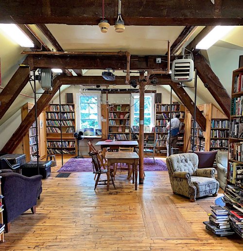 Cozy library interior with wooden beams, bookshelves, a table, and a person organizing books.