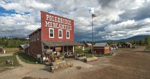 A rustic red mercantile building with a flag, surrounded by trees and mountains under a cloudy sky.