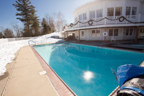 A clear blue pool surrounded by snow, with a white building and trees in the background on a sunny day.