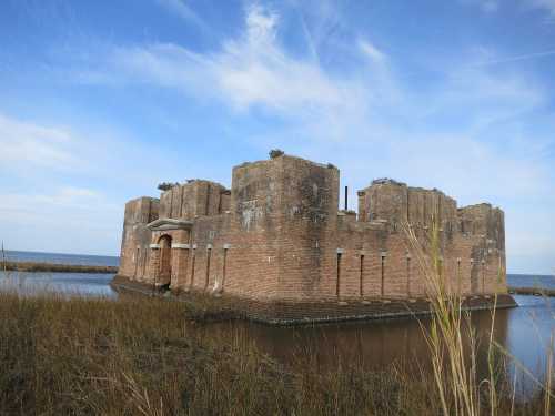 A weathered brick ruin surrounded by water and tall grass, under a blue sky with scattered clouds.